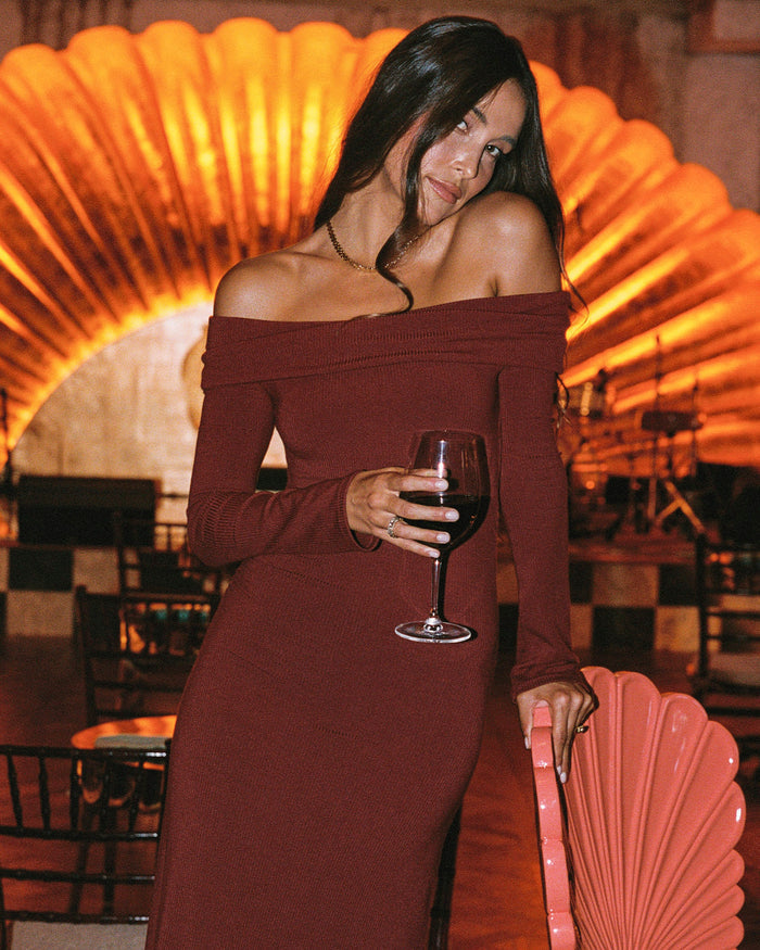 Lifestyle front view of a model wearing the LSPACE Carrie Dress in sangria, featuring an off-the-shoulder folded neckline and body-hugging maxi silhouette in ribbed sangria knit fabric, photographed in an atmospheric venue with a glowing fan-shaped backdrop.