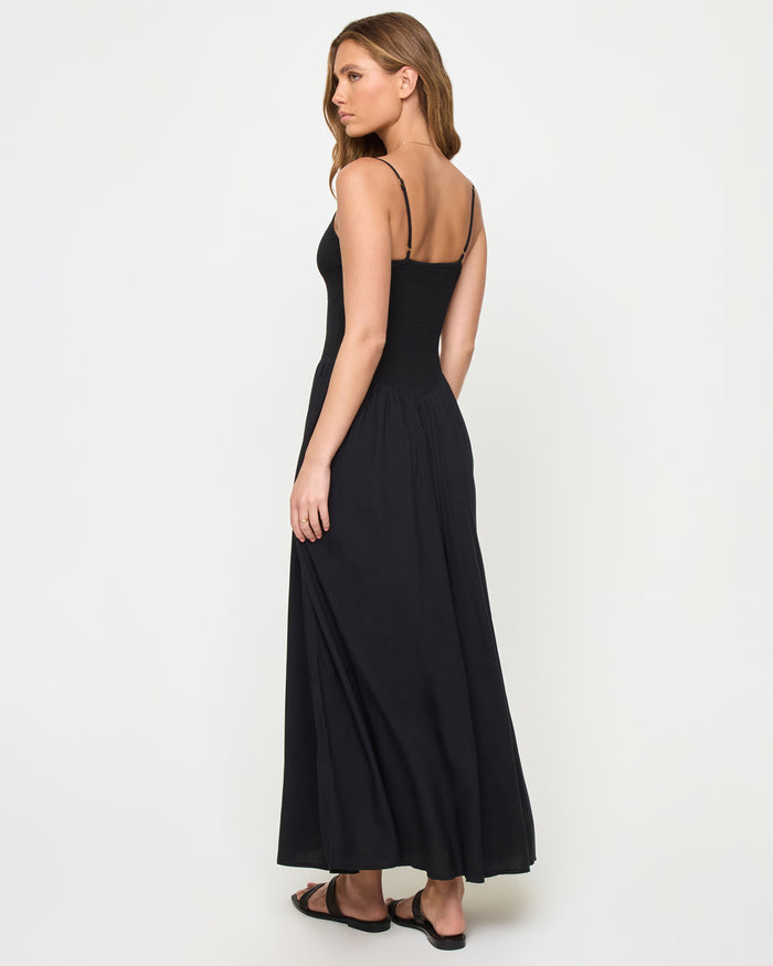 Three-quarter back view of a model wearing the LSPACE Misha Midi in black, showing thin spaghetti straps, an open back, and a full flowing black skirt.
