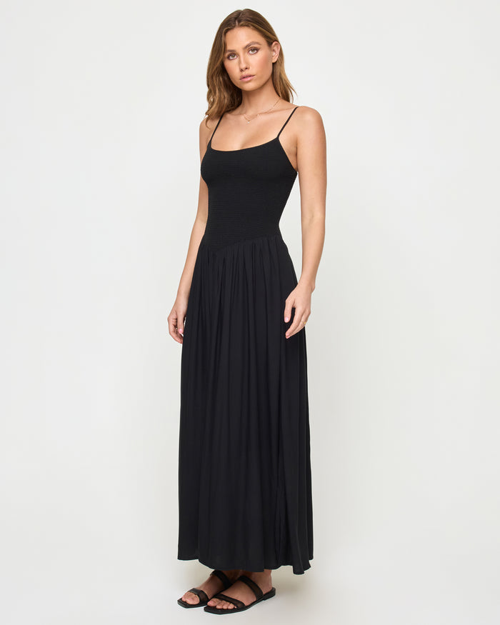 Three-quarter view of a model wearing the LSPACE Misha Midi in black, featuring thin spaghetti straps, a scoop neckline, a smocked fitted bodice, and a full flowing black skirt.