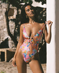 Front view of a model wearing the LSPACE flower play Piper One Piece Swimsuit with a deep V-neckline and tie-front detail, leaning against a pillar outdoors.