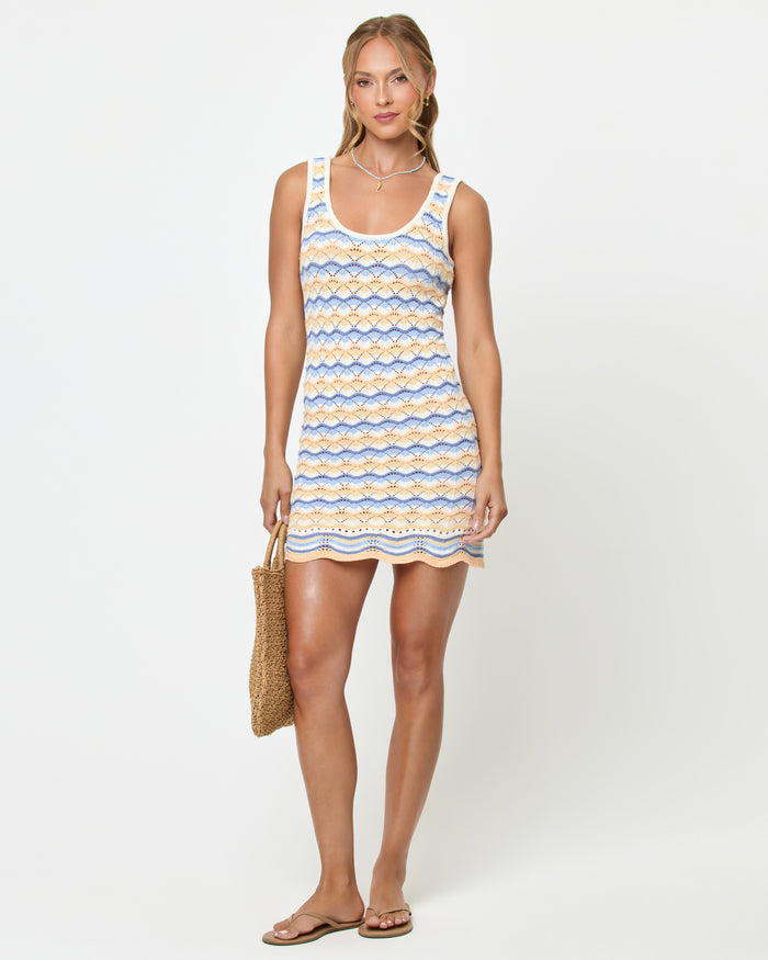 Front view on model of the LSPACE Sunchaser Dress in Daydreamer, a sleeveless crochet mini dress in a blue, peach, and white scallop pattern with scalloped hem, styled with flat sandals.