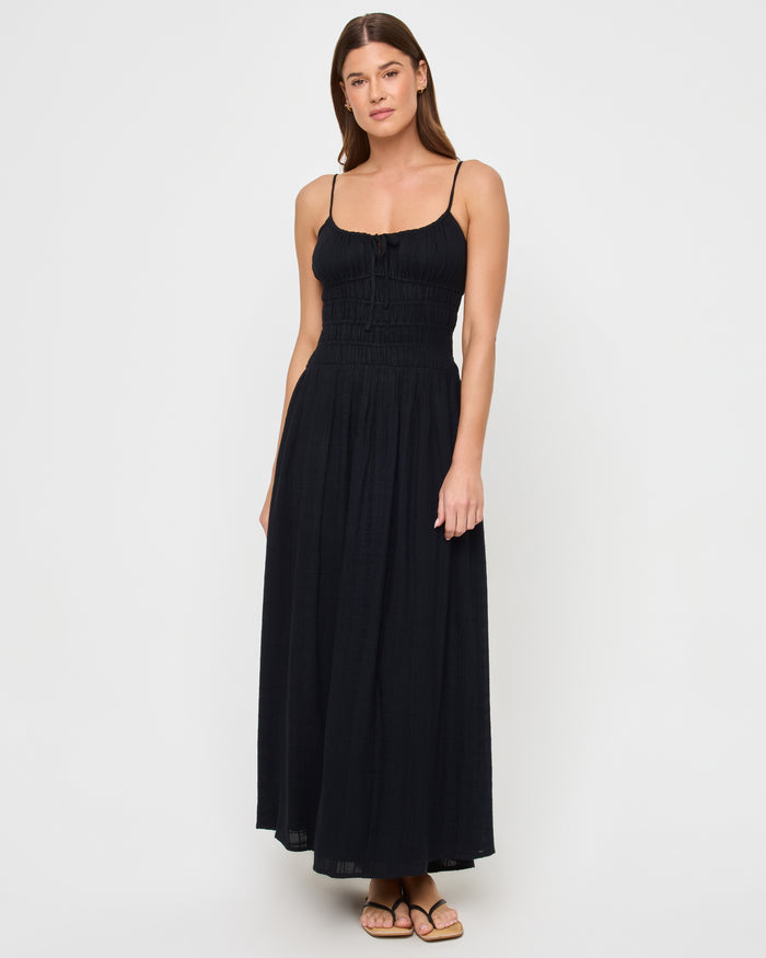 Front view of a model wearing the LSPACE Tala Dress in black, a full-length maxi dress with thin spaghetti straps, a smocked bodice with a front center tie detail, and a flowing skirt.