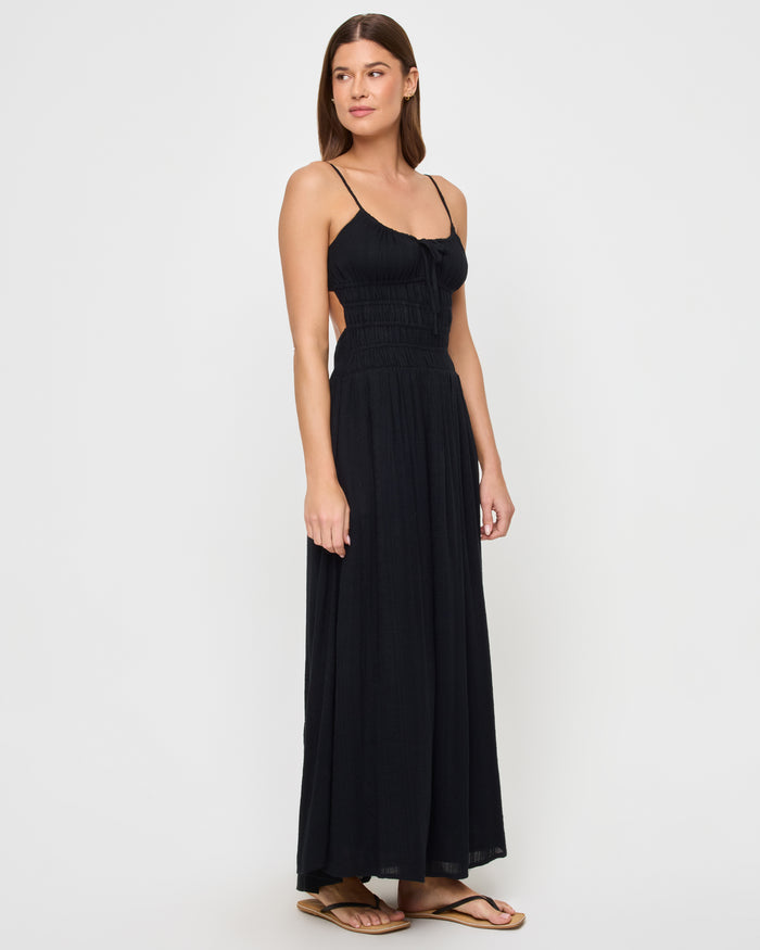 Angled view of a model wearing the LSPACE Tala Dress in black, showing the open-back cutout design with spaghetti straps and a smocked waistband on the flowing maxi skirt.